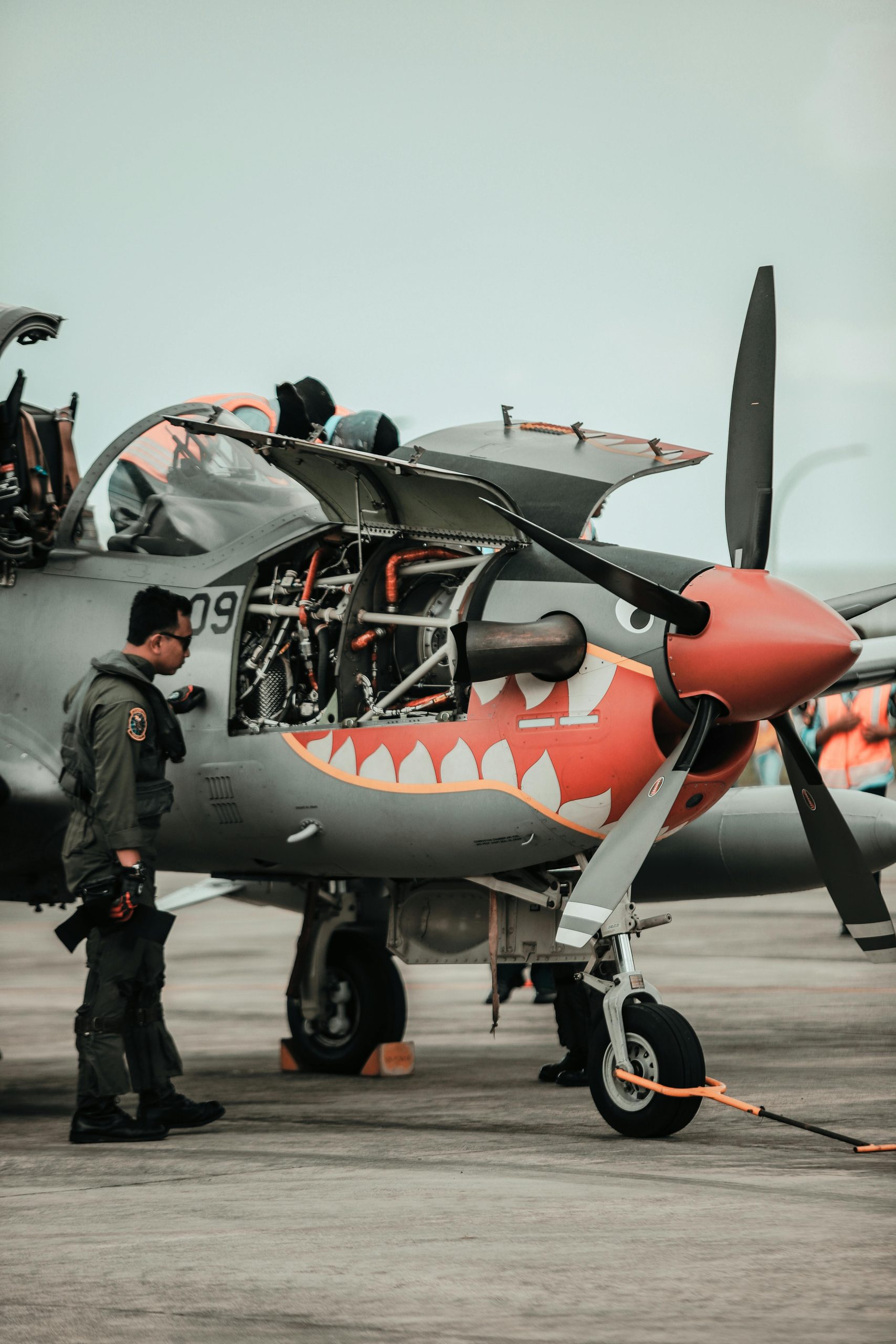 A pilot inspects a military aircraft with an open engine on the tarmac.